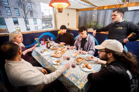 Five diners sit around a small table, as a man stands looking on