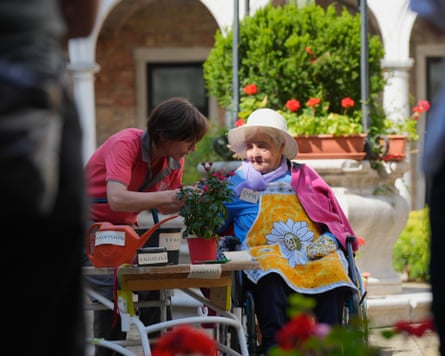 A carer speaks with an elderly woman in a hat and apron sitting in a courtyard near a flowering bush
