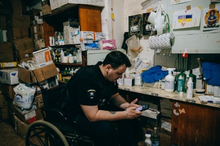 A man looks at his phone while sitting in a wheelchair in a room packed with medical supplies