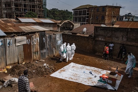 Four people in white hazmat suits carry a body out of a clearing in a shantytown as others look on