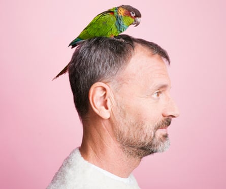 Close up of side view of Tim Dowling with a parrot on his head, against pink background