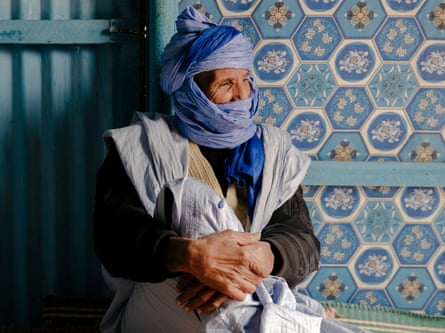 A barefoot man in a scarf covering his head and face sits on a mat and leans against a wall of corrugated iron and decorative tiles.