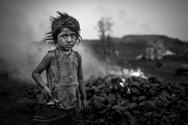 A child blackened with coal dust stands in front of a smouldering pile of coal