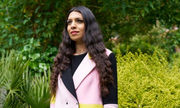 Faiza Shaheen wearing a pink, yellow and black jacket and looking into the distance, with lots of plants and vegetation behind.