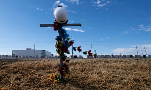 A metal cross on the side of the road is covered with flowers and topped with a worker's helmet.