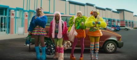 Four young women in brightly coloured clothing, wigs, fake fur coats, fluffy boots and coloured tights or socks stand in front of an orange vintage car.