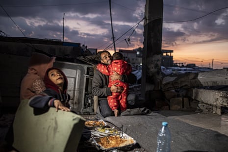 Palestinian Muhammad al-Durra sits on the floor with his children on Wednesday in Rafah. Plates of food are placed between them and al-Durra is holding a toddler in a red babygrow.