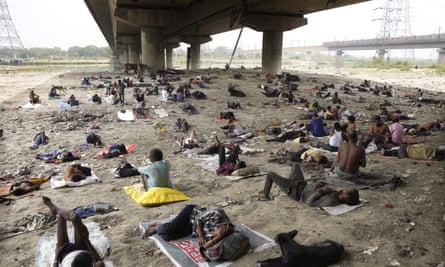 Dozens of Indian people with a few belongings lying on mats on bare earth in the shade under a motorway flyover