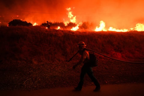 a firefighter carries a hose along a road as a fire rages in the background