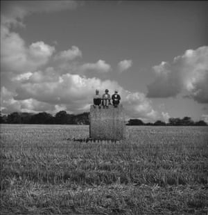 Three men sitting atop a tall hay bale, which stands in a ploughed field.