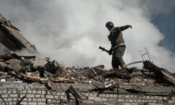 A man in a firefighter's uniform holds a hose while standing on top of the rubble of a building with smoke billowing in the sky behind