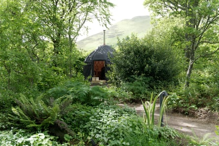 A yurt with a silver chimney among trees and shrubs with hills behind