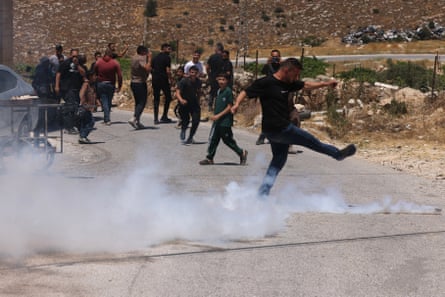 A young man dressed in black kicks a teargas cylinder with a cloud of white smoke-like gas streaming around his feet, as others stand behind him on a village road and look on.