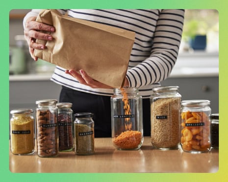 A woman pours dried food from a paper bag into a glass jar among labeled pantry jars