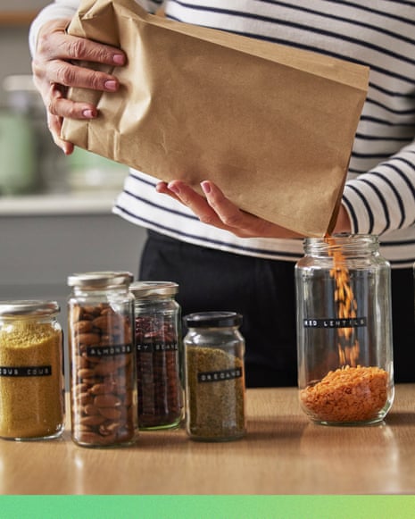 A woman pours dried food from a paper bag into a glass jar among labeled pantry jars