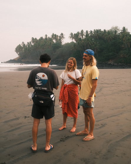 Three people stand on the sand, one wearing a black Save the Waves t-shirt.