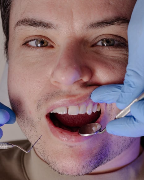 A dentist wearing blue gloves examines a patient's open mouth with dental instruments