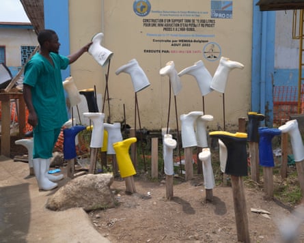 A healthcare worker in green scrubs and protective boots holds a white rubber boot beside rows of boots mounted on wooden stakes outside a medical facility