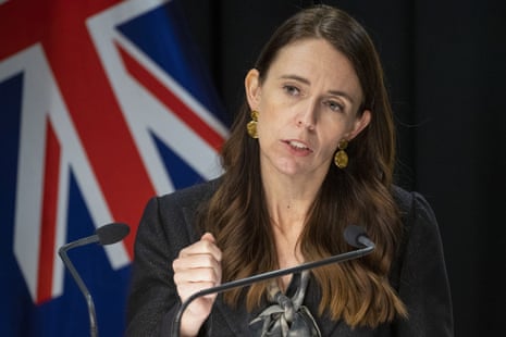 Jacinda Ardern looking serious as she speaks in front of a New Zealand flag