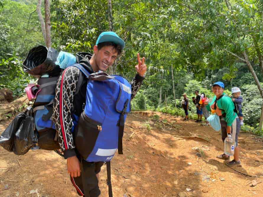 A man carrying two backpacks waves at the camera while a woman in the background  carries a baby on her back