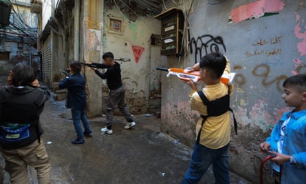 Five young children play in a narrow street. Three hold up plastic machine guns. On one of the walls is a spray-painted red triangle. 