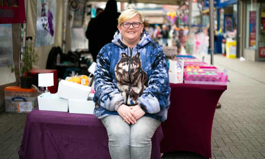 Woman sits on edge of her market stall, collection of boxes behind her, wearing zip-up jacket with picture of husky dog