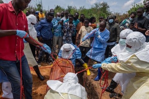 Red Cross workers in protective gear lower a body bag into a grave using ropes, surrounded by onlookers