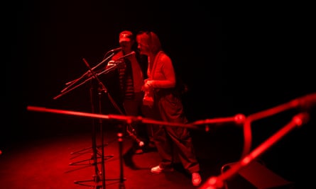In a red-lit room, two people lean in towards microphones. 