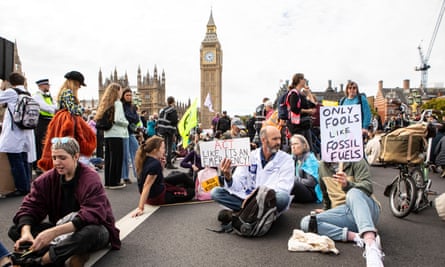 A crowd sitting on Westminster Bridge holding signs like “Only fools like fossil fuels” with Big Ben in the background