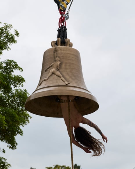 A person hangs upside down through a large suspended bell, their long hair falling downward