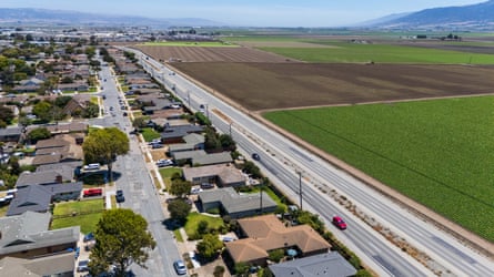 Aerial view of four-lane highway dividing single-family homes on one side, and flat fields on the other, with hills in the distance.