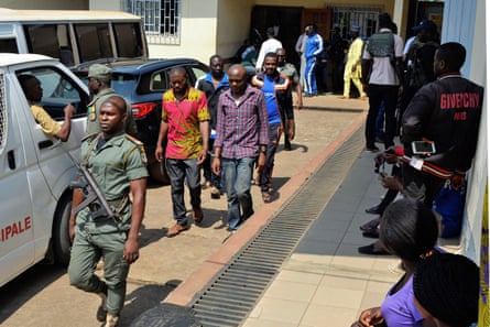 Four men walk down a street outside a building, led by an armed guard