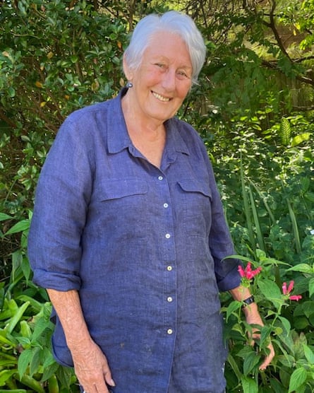 A woman in a long blue shirt, smiling, trees and plants behind her
