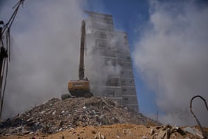 A large crane on a pile of rubble is framed by clouds of dust with a badly damaged building in the background