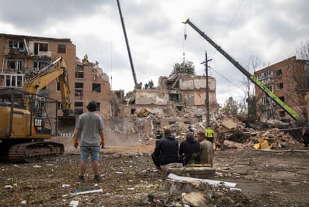 Men sitting and standing, looking at the destroyed building, with emergency workers and cranes