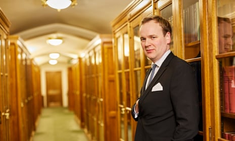 William Wragg in the Palace of Westminster, 18 December 2022: he is standing to the side in a corridor lined with glass-fronted wooden cabinets that are filled with books. He wears a dark suit jacket with white shirt and tie, and is holding one hand inside his jacket.