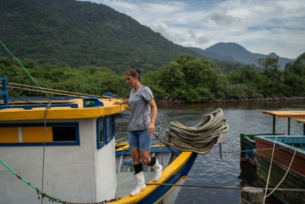 Uma mulher de meia-idade, de shorts jeans e botas de borracha brancas, caminha ao longo das bordas de um barco de pesca, com colinas verdes ao fundo
