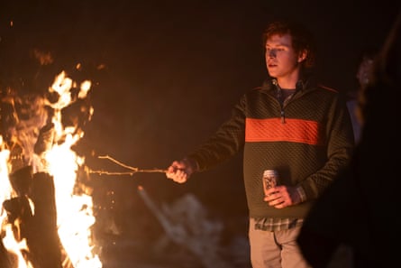 Holding a can of beer, the boy puts a stick to the flames of a bonfire