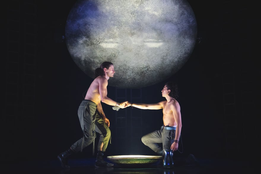 Stephen Madsen et Carl Richmond, les acteurs qui jouent Achille et Patrocle, sur scène.  Ils se font face, la main droite reliée au-dessus d'une grande lumière.
