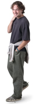 A young man with curly brown hair wearing a grey T-shirt, trousers and trainers, with a sweatshirt tied round his waist