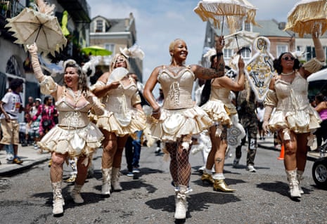 Dancers in a street wear ivory-coloured bustier and corset tops, flared skirts and boots; some hold matching parasols and fans