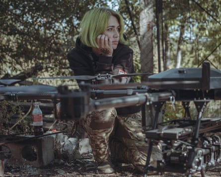 A woman with green bobbed hair wearing a black hoodie and combat trousers leans on a black drone