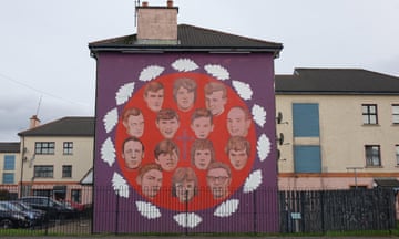 A mural on the side of a building in Bogside, Derry, commemorates the unarmed civilian protesters shot dead by British soldiers on Bloody Sunday (30 January 1972): their faces and a cross are depicted on a bright red circle, framed by white leaves and on a darker purple background which stretches across the whole wall.