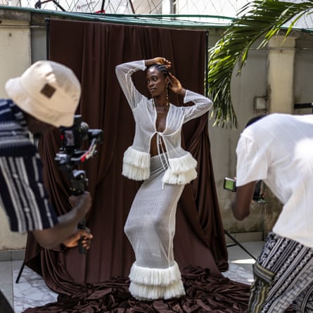 A model poses for photographers in front of a hut; she wears a white top and tight, long skirt with thick fringing around the hem.
