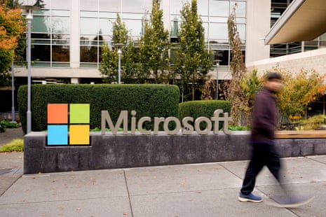 A blurred man walks past a Microsoft sign and logo on its campus, surrounded by hedges
