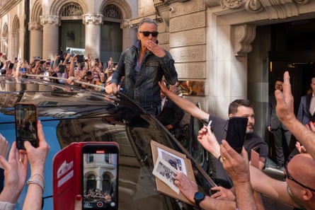 Bruce Springsteen blows a kiss while standing on the side of a vehicle surrounded by fans