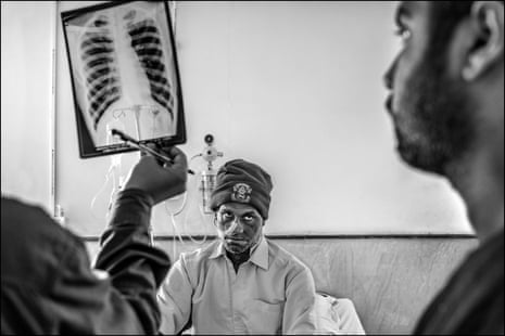 Dois médicos examinam um raio-X enquanto o paciente está sentado em uma cama com um gorro de lã e um tubo saindo do nariz