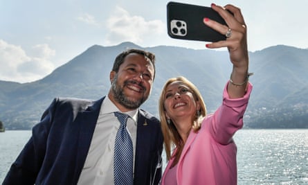 Giorgia Meloni takes a selfie with Matteo Salvini, both wearing suits, in front of a mountain-surrounded lake during a conference in Italy
