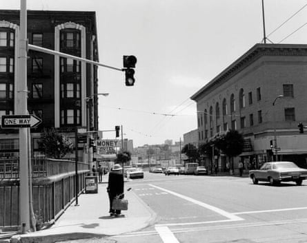 A black-and-white image of a city corner, with brick three- and four-story buildings and a person waiting to cross the street.