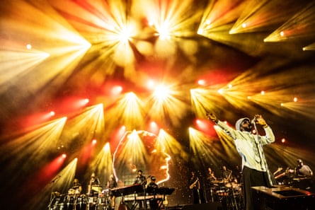 A young black African man wearing sunglasses and a cap performs on stage with a large band behind him and lots of lighting effects